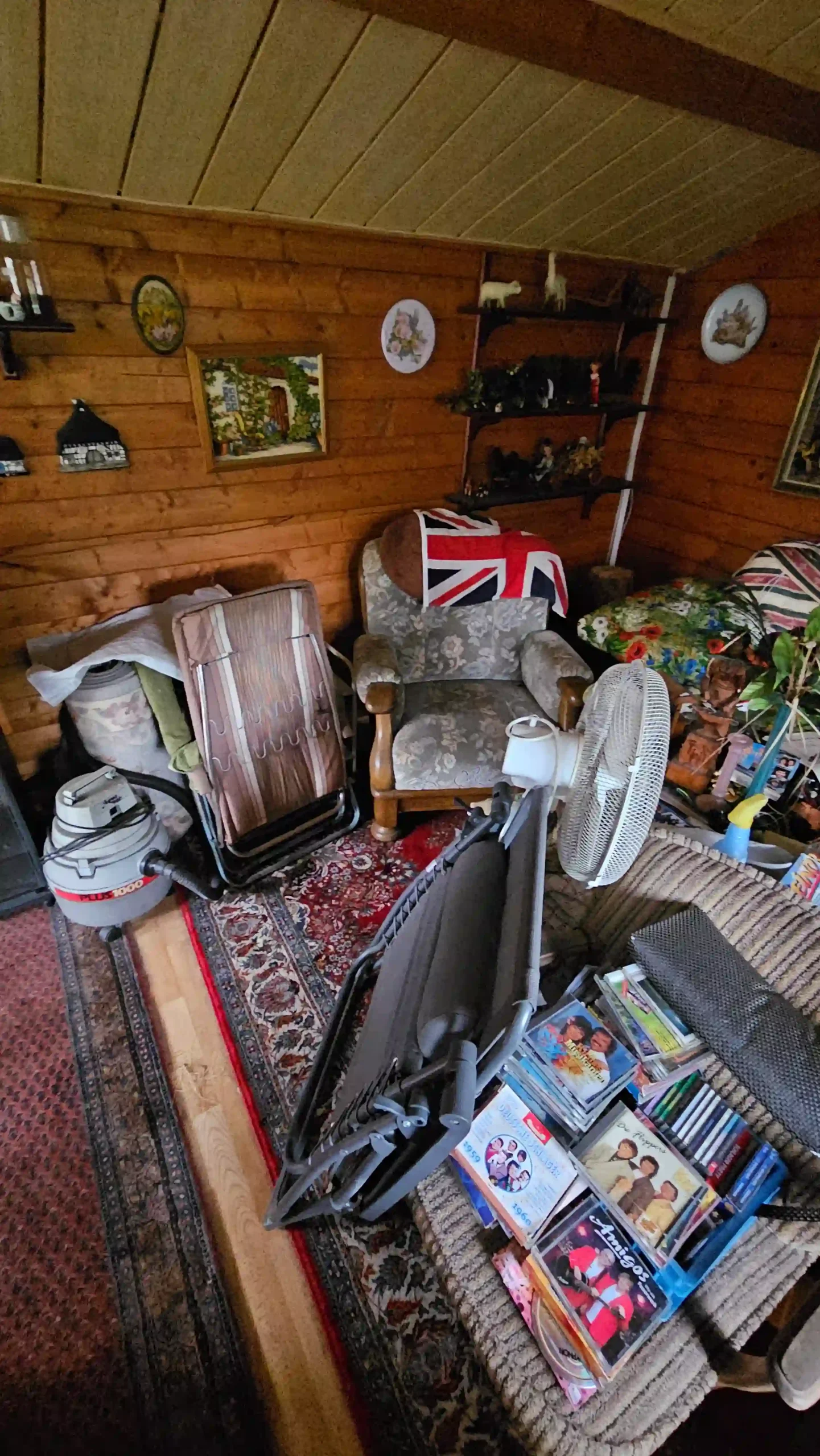 Haushaltsauflösung Raum unter einer Holz-Dachschräge mit Sessel, Klappbett, Ventilator, Staubsauger und gelagerten Gegenständen